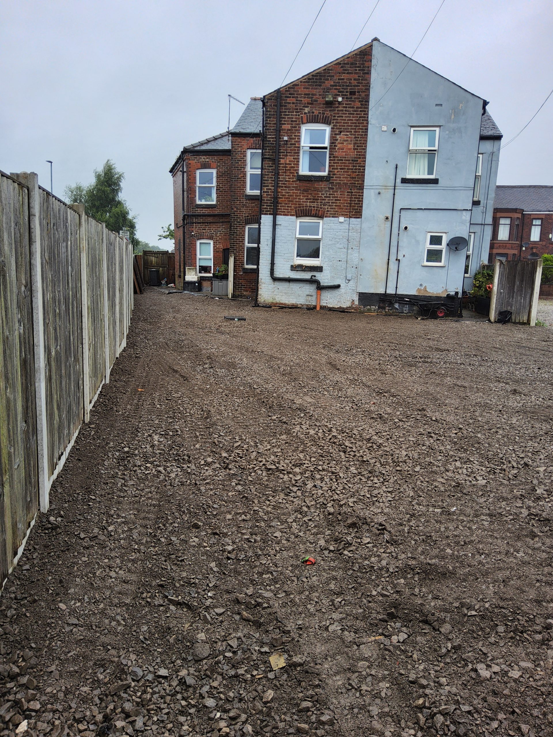 The completed mixed-use development site showing cleared, leveled crushed-stone ground adjacent to the two-story brick and blue-rendered residential building, with a wooden fence along the boundary under an overcast sky